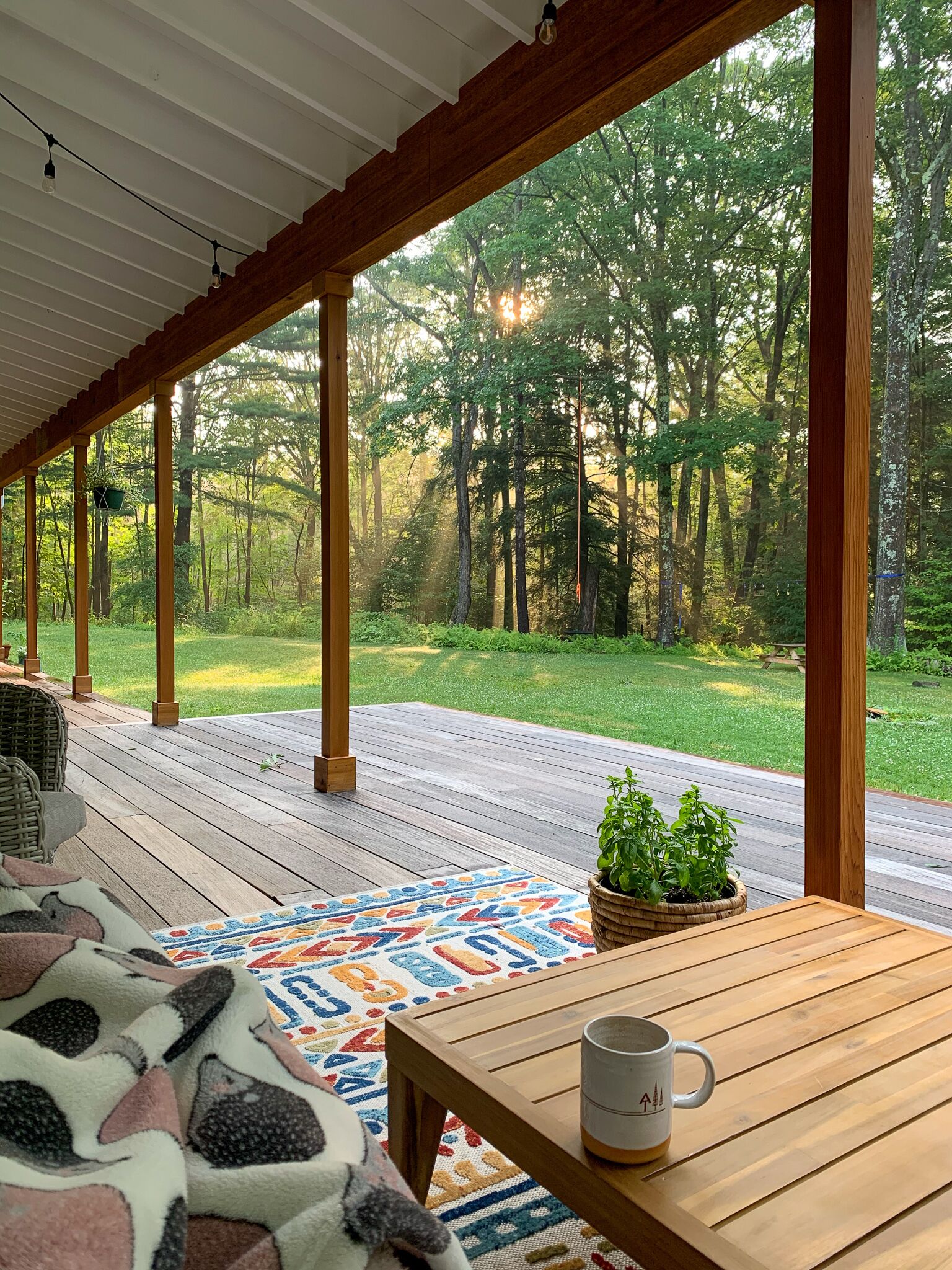 A covered outdoor deck with a wooden table and a mug, overlooking a forested area.