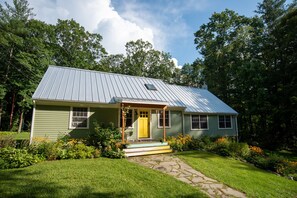 A single-story house with a green exterior and a yellow front door, surrounded by a well-maintained garden and a stone pathway leading to the entrance. - Secluded Family Home in Serene Stockbridge Woods (Stockbridge)
