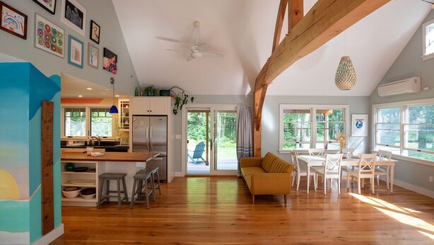 A spacious living area with a wooden ceiling, a kitchen with a refrigerator, and a dining area with white chairs.