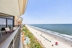 A balcony with a view of a beach, a pool, and a clear sky.