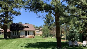A two-story house with a well-maintained lawn, a patio with outdoor furniture, and a large tree in the foreground. - Beautiful river home with large yard, sitting area and fire pit! (Estes Park)