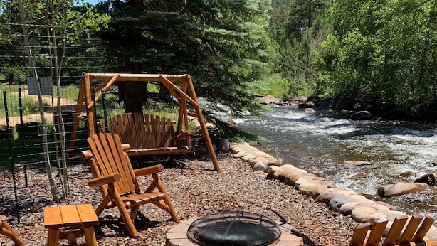 A wooden deck with Adirondack chairs and a fire pit, overlooking a river. - Beautiful river home with large yard, sitting area and fire pit! (Estes Park)