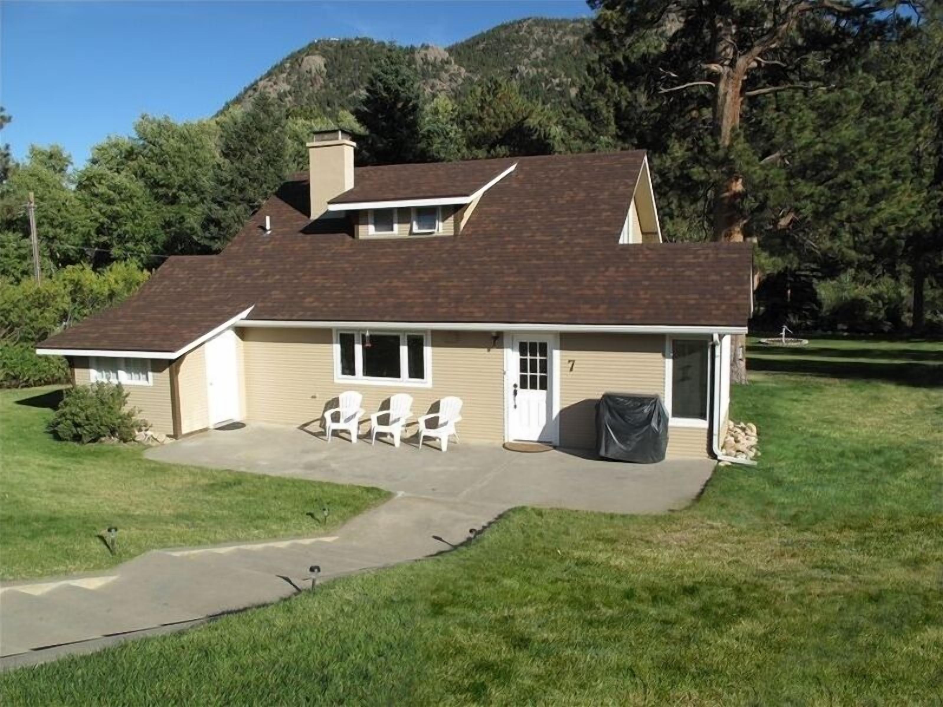 Une maison de plain-pied avec un toit brun, un revêtement beige et un patio couvert équipé de trois chaises blanches.