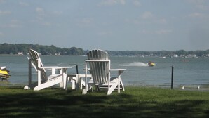 A lakeside setting with white Adirondack chairs and a table, a jet ski in the water, and a distant shoreline with buildings. - Macatawa Lake View Cottage by State Park and Bike Trails (Holland)