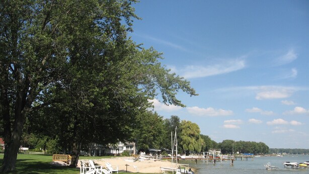 A lakeside park with a grassy area, white chairs, and a sandy beach. - Macatawa Lake View Cottage by State Park and Bike Trails (Holland)