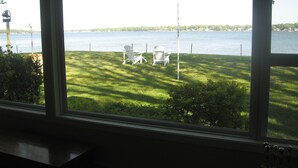 A view from a window overlooking a grassy area with two white chairs and a table, a body of water, and a distant shoreline with trees and buildings. - Macatawa Lake View Cottage by State Park and Bike Trails (Holland)