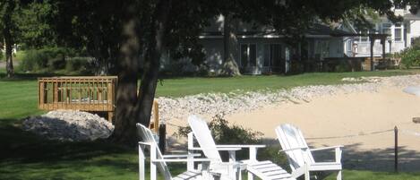 Zona de playa con sillas reclinables blancas y un banco de madera cerca de un árbol, con una casa y la playa al fondo.