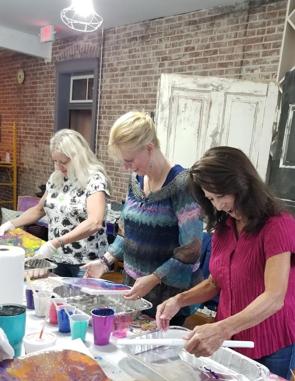 Three people working at a table with art supplies, in a room with brick walls and a white door.