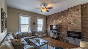 A living room with a brick fireplace, a ceiling fan, a sectional sofa, and a television. -  SPACIOUS HISTORIC DOWNTOWN LOFT!  BEST LOCATION IN THE HEART OF TOWN! (Hermann)