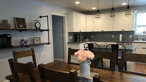 Un comedor con una mesa y sillas de madera, un jarrón con flores, y una cocina al fondo con armarios blancos y salpicadero decorado.