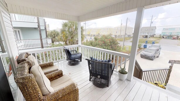 A covered porch with wicker furniture, a potted plant, and a view of a street with buildings and a car. -     Sun, Sand & Serenity — Your Gulf Getaway Awaits (Gulf Shores)