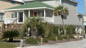 A two-story house with a green roof, a front porch, and a small garden with palm trees and shrubs. -     Sun, Sand & Serenity — Your Gulf Getaway Awaits (Gulf Shores)