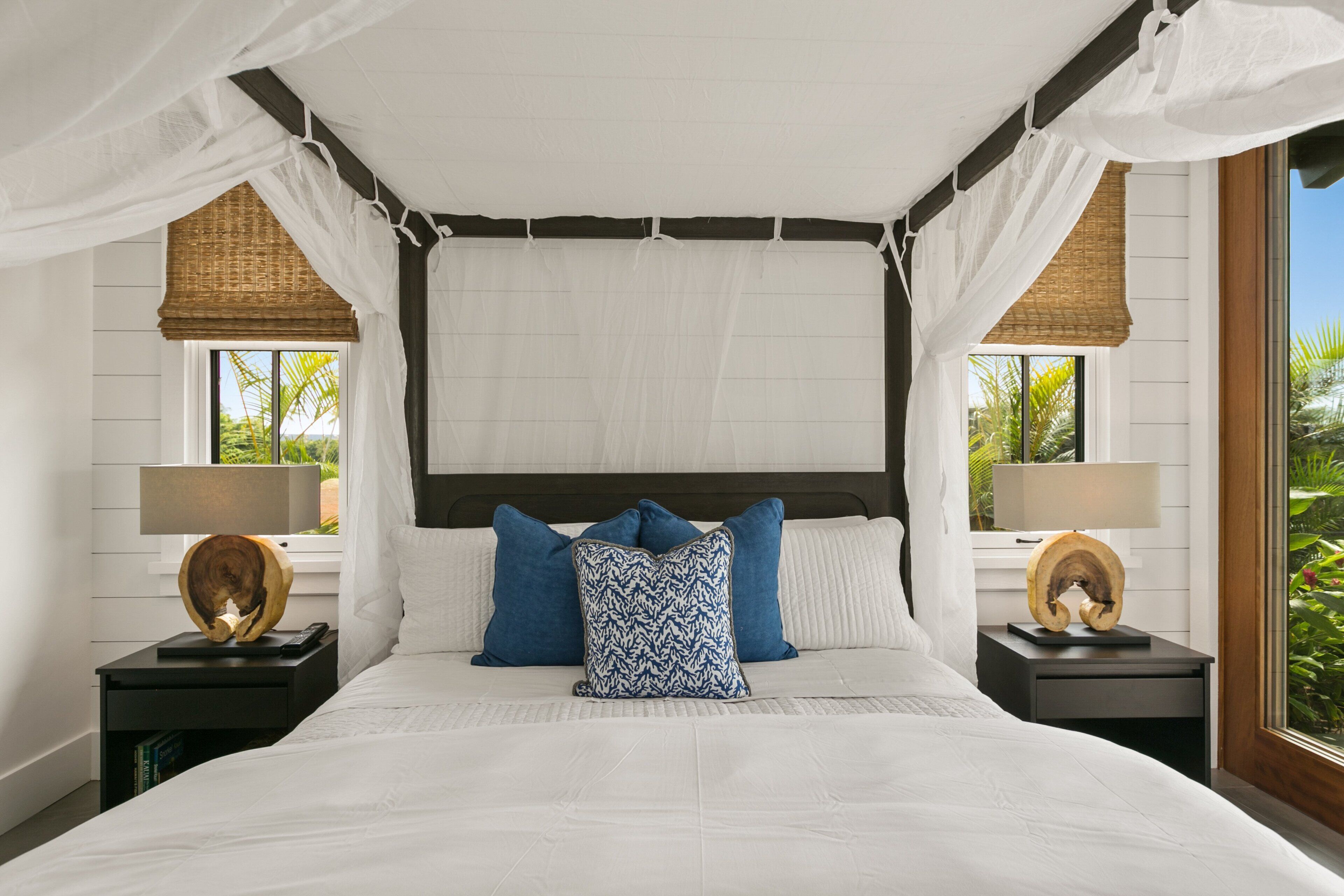 A bedroom with a canopy bed, two bedside tables with lamps, and a view of greenery outside.