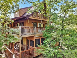 Une cabane en bois à deux étages, dotée d’un balcon à chaque niveau, et entourée d’une végétation luxuriante.