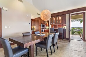Un comedor moderno con mesa de madera y sillas de cuero, una cocina con armarios de madera, y vista al océano a través de un ventanal abierto.