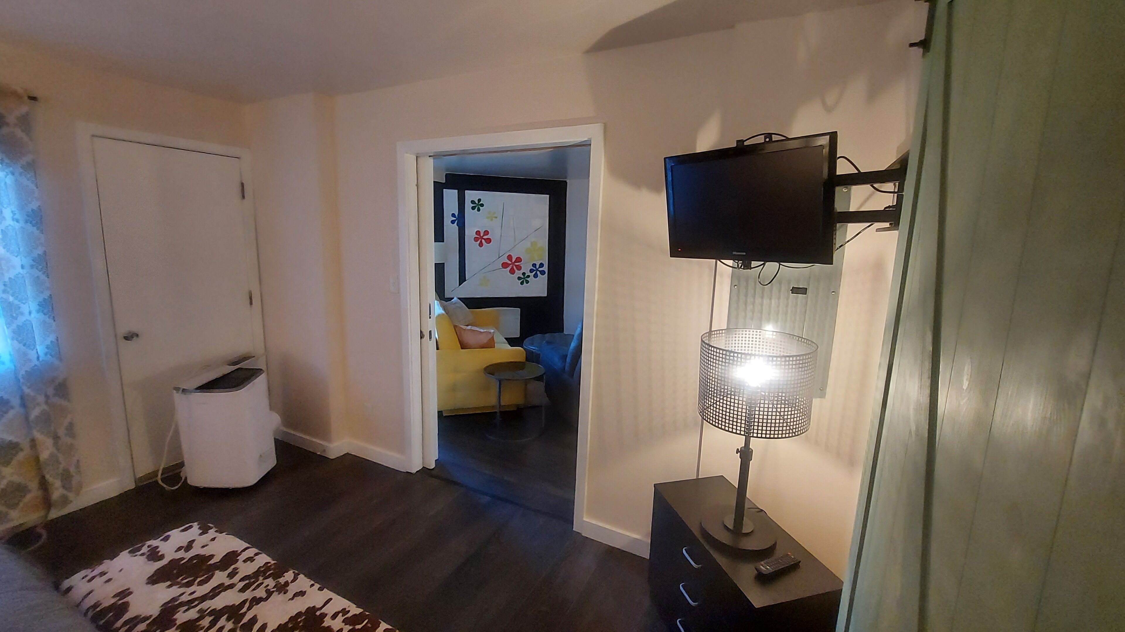 Et værelse med fjernsyn ophængt på væggen, et lille bord med lampe, en sofa og en tæppe med mønster.