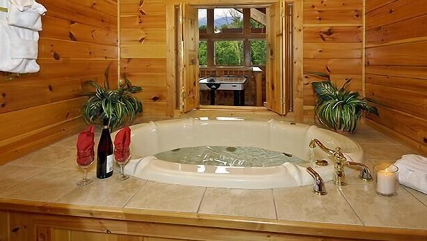 Ein Badezimmer einer Holzhütte mit einer freistehenden Badewanne, zwei Zimmerpflanzen, einem Handtuchhalter und einem Fenster zu einem Außenbereich.
