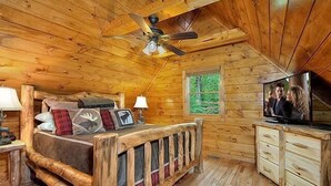 Un dormitorio tipo cabaña de madera con una cama, un ventilador de techo, un televisor y una ventana con vistas a follaje.
