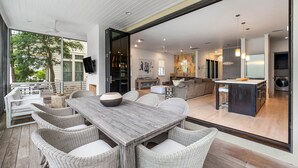 An open-plan living area with a dining table and wicker chairs, a kitchen, and a view of the outdoors. - Blissful days, barefoot nights at Beach Bliss / Private Pool / Gulf Front Beach Club / 6 Bikes (Santa Rosa Beach)