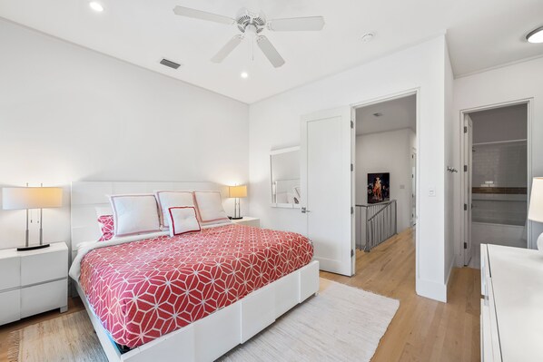 Un dormitorio con cama, mesitas de noche blancas, ventilador de techo y puerta que da al baño.