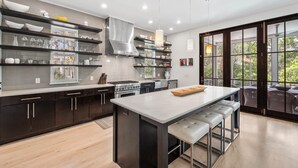 A modern kitchen with dark cabinetry, a central island with stools, and a large window offering a view of greenery. - Blissful days, barefoot nights at Beach Bliss / Private Pool / Gulf Front Beach Club / 6 Bikes (Santa Rosa Beach)