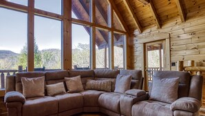 A wooden cabin interior with a large sectional sofa, large windows, and a view of the outdoors. - Mountain View Gameroom-hot tub-outdoor pool access free wifi pet friendly & more (SEVIERVILLE)