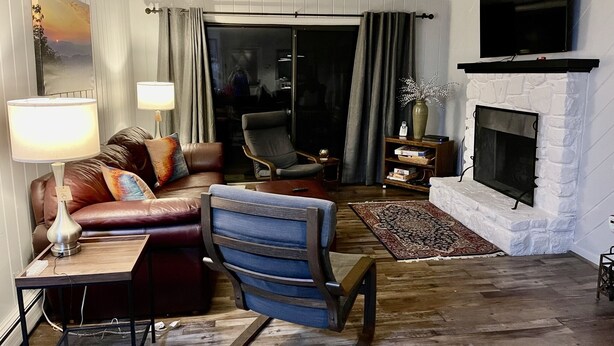 A living room with a fireplace, a sofa, a chair, a table with a lamp, and a large window. - BEECH MOUNTAIN HIGH - WALK TO SLOPES! (Beech Mountain, NC)