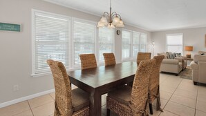 A dining area with a dark wooden table and wicker chairs, large windows with blinds, a chandelier, and a living area with a sofa and lamp. -  Private Beach Access l CONVENIENCE: 2 minute walk to the Beach! (Santa Rosa Beach)