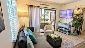 A living room with a sofa, armchairs, a coffee table, a TV, a plant, and a sliding glass door leading to a balcony.