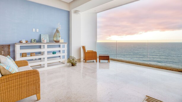 A modern living room with a view of the ocean at sunset, featuring a white console table, wicker furniture, and a potted plant. - Luxury Oceanfront Condo with unbeatable views (Puerto Vallarta)