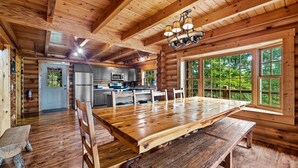 Interior de una cabaña de madera con una gran mesa de comedor, bancos de madera, una zona de cocina y ventanas amplias con vistas al bosque.