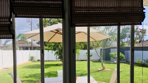 Un patio con mesa y sillas, un área cubierta con sombrilla y vista a un jardín trasero con cerco y árboles.
