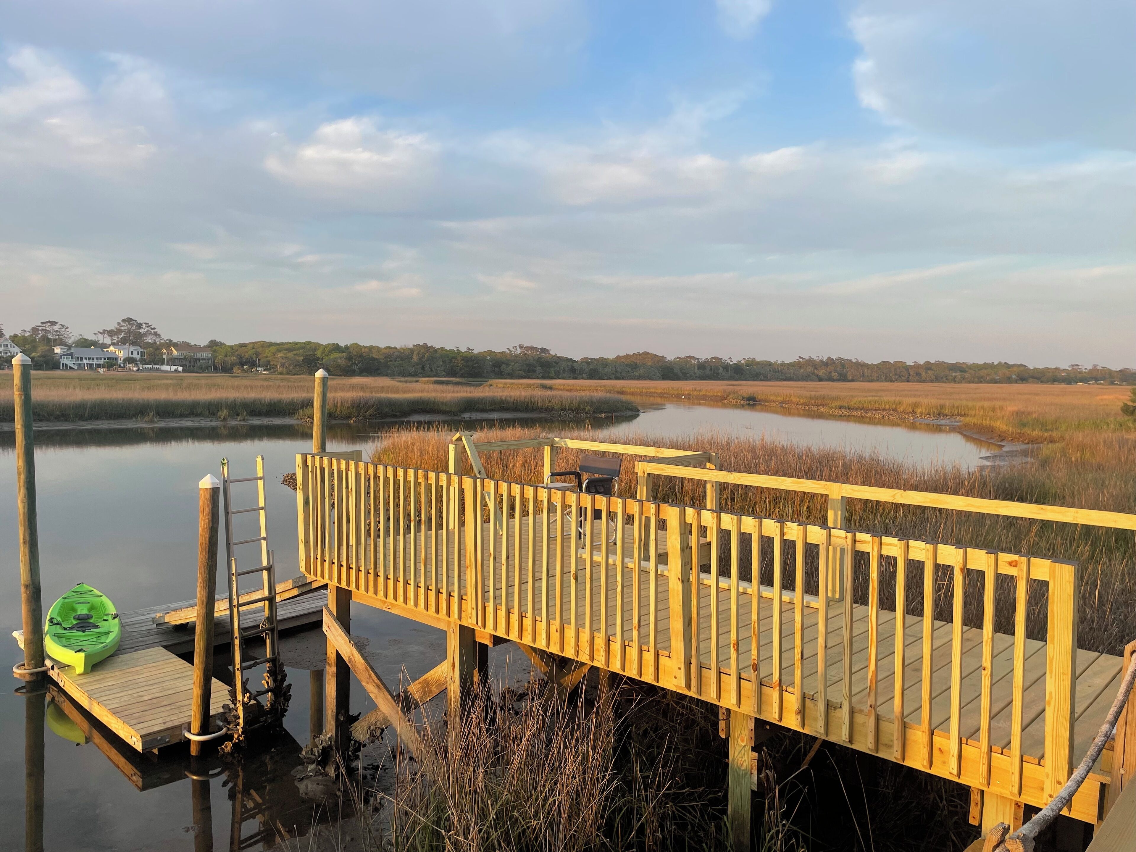 A wooden dock extending over calm water, surrounded by marshland and distant houses.