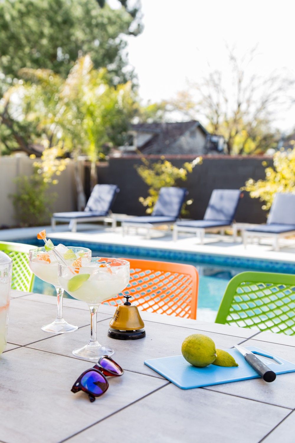 A poolside table with drinks, a bell, a cutting board, and sunglasses.