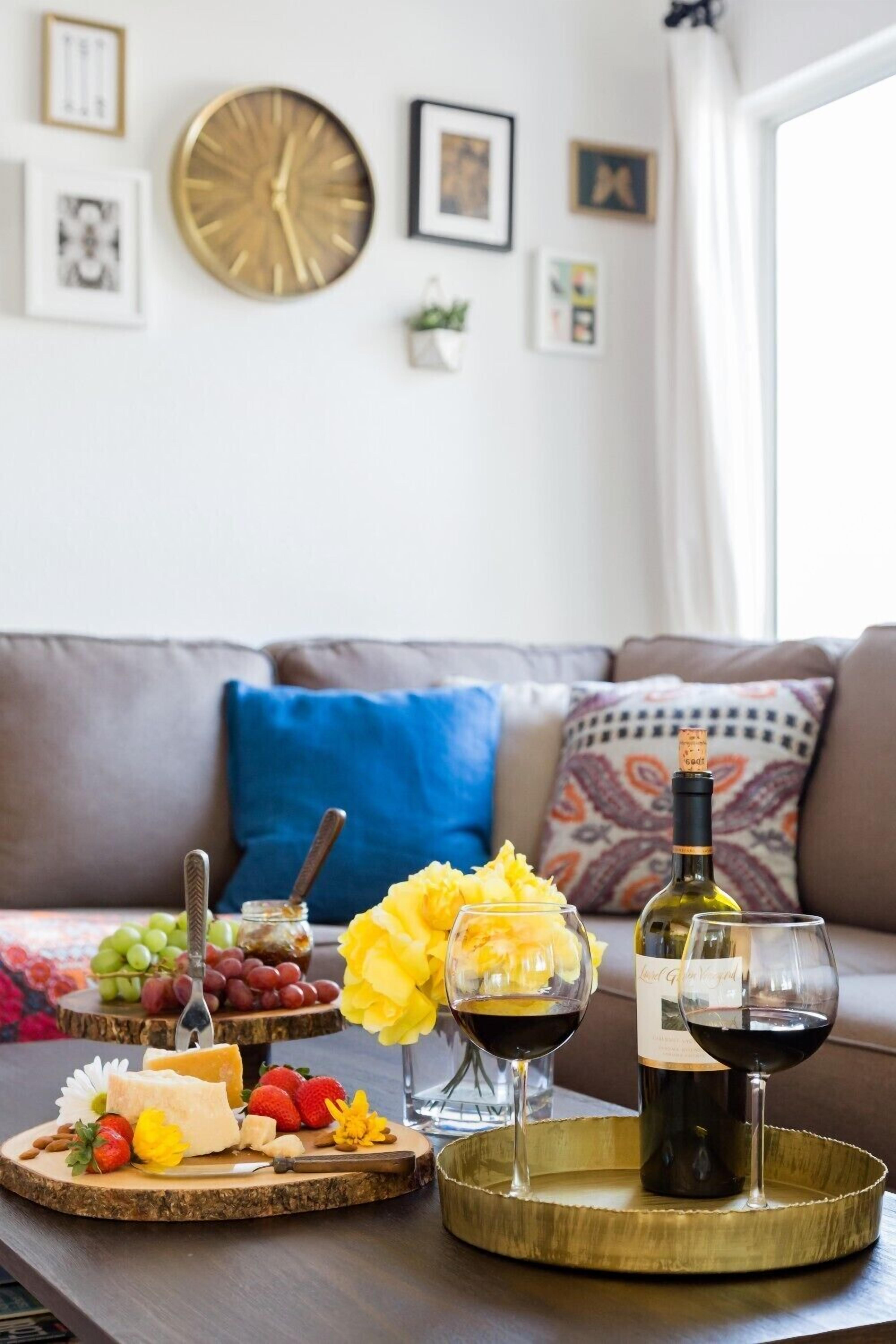 Uma mesa de jantar com um prato de queijos, uvas e copos de vinho, numa sala de estar com um sofá e almofadas decorativas.