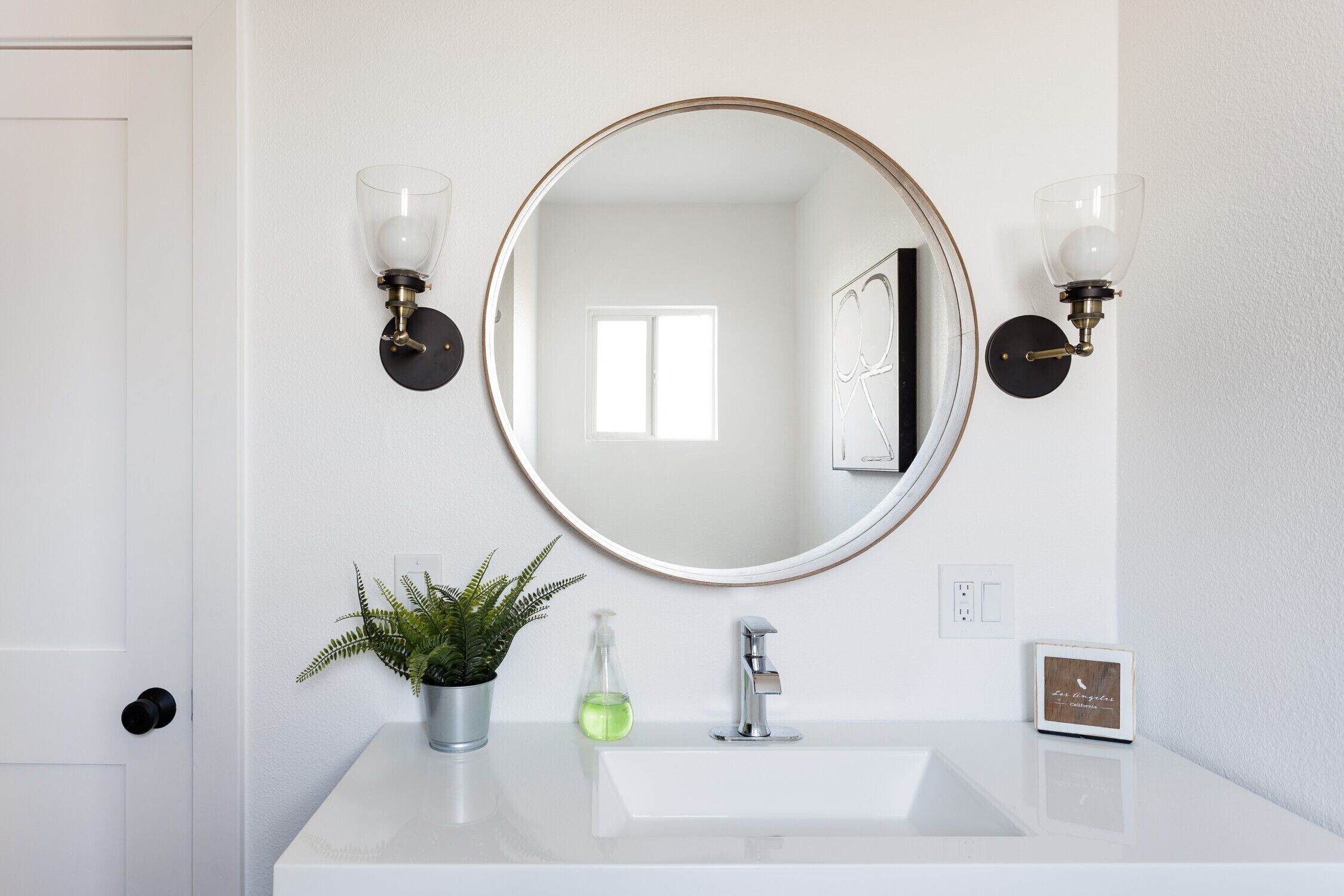 Une salle de bain avec un lavabo blanc, un miroir rond, deux appliques murales, une plante en pot, un distributeur de savon et un tableau encadré.