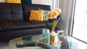 Un salon moderne avec un canapé en cuir foncé, des coussins aux motifs jaunes et bleus, et une table basse en verre agrémentée d’objets décoratifs, notamment une étoile de mer et un vase avec des fleurs jaunes.