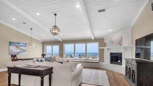 A spacious living room with a large white sectional sofa, a dark wooden coffee table, and a fireplace. There are large windows offering a view of the ocean. - My Island Pad: Beachfront Home, Fully Renovated (Pensacola Beach)