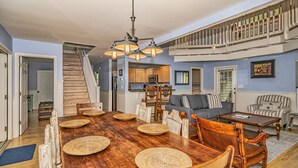 A spacious living area with a wooden dining table, a staircase leading to an upper level, and a kitchen in the background.