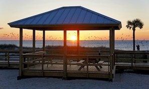 En træpavillon med ståltag, beliggende på en sandstrand med udsigt til solnedgangen over havet.
