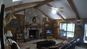 A cozy cabin interior with a stone fireplace, wooden beams, a ceiling fan, and a large window.