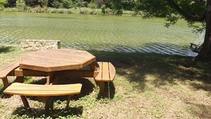 A hexagonal wooden picnic table with benches, situated by a lake with a stone structure in the background.