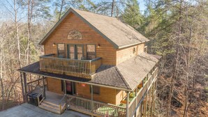 A two-story log cabin with a balcony and a deck, surrounded by trees. - Outdoor pool access, Private putt putt-Close to Pigeon Forge-outdoor kitchen-game room and  more.... (PIGEON)