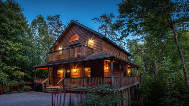 A two-story wooden cabin with a balcony, surrounded by trees. - Outdoor pool access, Private putt putt-Close to Pigeon Forge-outdoor kitchen-game room and  more.... (PIGEON)