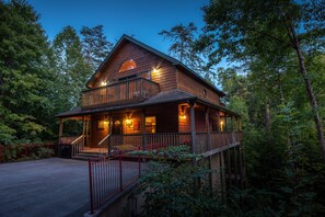 A two-story wooden cabin with a balcony, surrounded by trees. - Outdoor pool access, Private putt putt-Close to Pigeon Forge-outdoor kitchen-game room and  more.... (PIGEON)