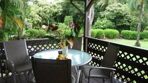 Een overdekt terras met een ronde tafel en stoelen, omgeven door een houten vlonder en weelderige beplanting.