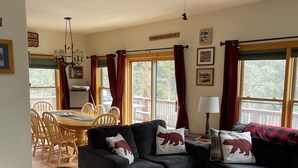Un salon confortable avec un ventilateur de plafond, un coin repas avec une table et des chaises, et de grandes fenêtres avec des rideaux.
