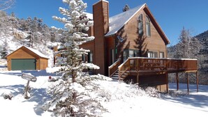 Une cabane en bois dotée d’une terrasse couverte, entourée d’arbres couverts de neige et surplombant un ciel bleu dégagé.