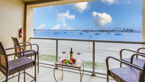 En balkong med flätade stolar, ett litet bord med en flaska och glas, och utsikt över havet med båtar.