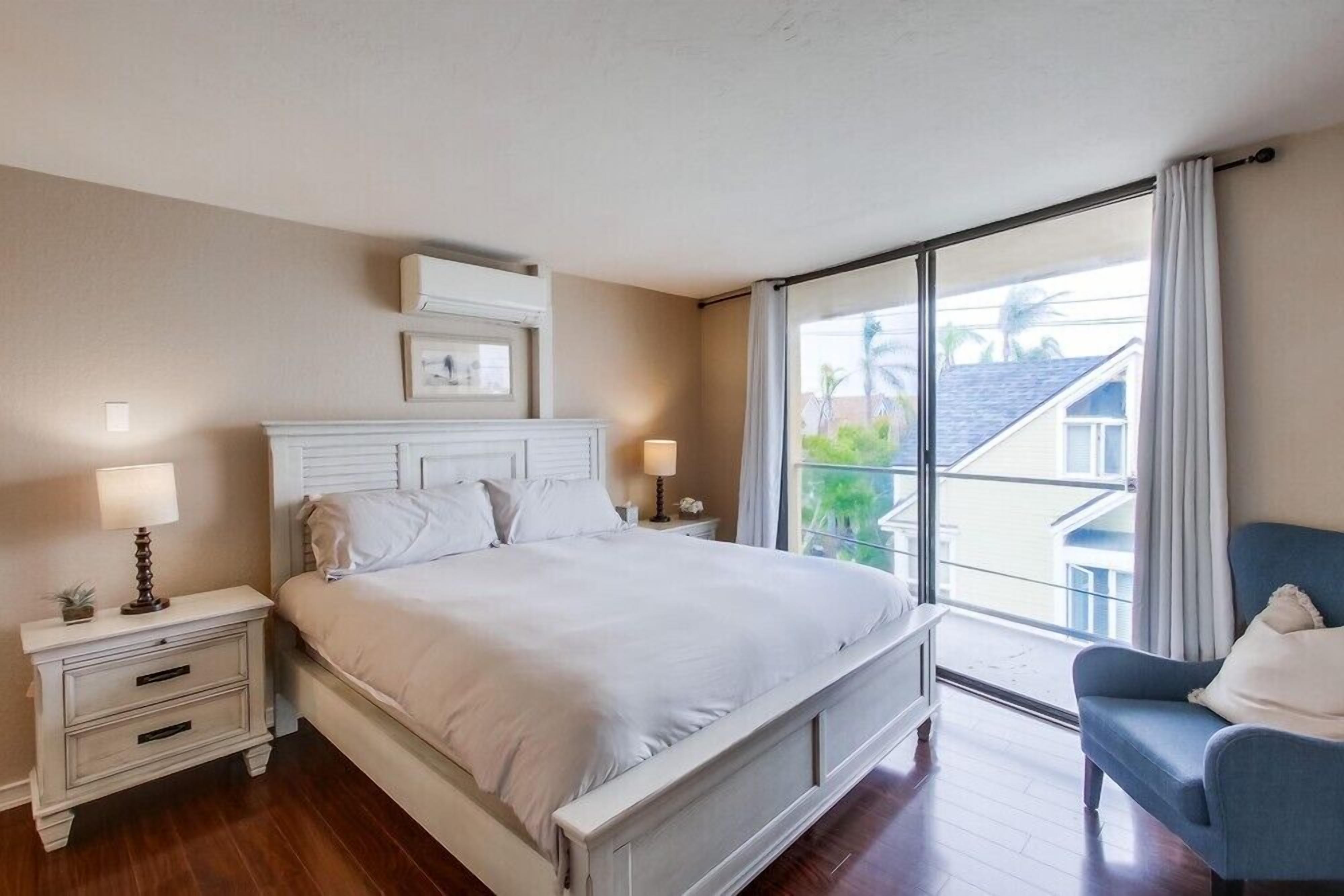 Un dormitorio con una cama grande, una mesita de noche con lámpara, una silla y vistas al exterior a través de un ventanal.